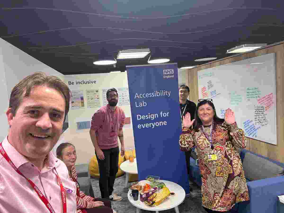 Chris, Danny, and members of the NHS England team smile for a selfie next to a roll up banner that reads "Accessibility lab - design for everyone"