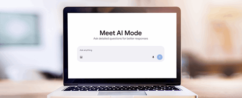 A laptop sits open on a wooden table indoors, displaying a screen with the heading “Meet AI Mode.” Below the heading is a prompt box that reads “Ask anything.” The room in the background is softly blurred, keeping the focus on the laptop’s screen.