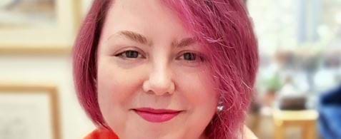 Claire is smiling at the camera. Her hair is a deep pink colour, and she is wearing a pink and orange shirt with a watercolour-esque design.