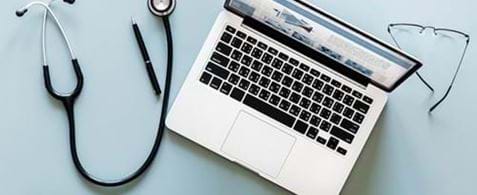 An open laptop on a table with a mobile phone, glasses, a pen and a stethoscope laying around it