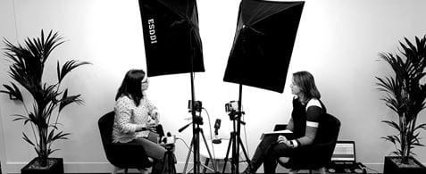 A black and white photo of two women sat facing across from each other. There is a dog at the feet of the woman on the left. There are camera lights shining on them and tall plants to the side of each of them.