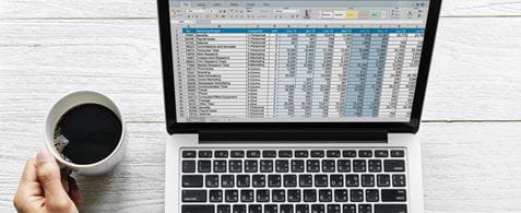 Hands on a trackpad of a laptop and holding a cup of coffee. There is a spreadsheet displaying on the trackpad.