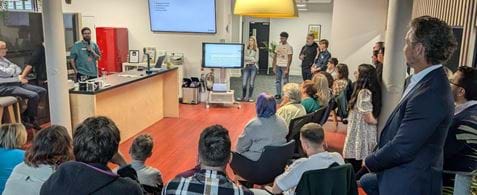 Danny speaks to a seated room filled with the Nexer team. They are sitting on stools and wooden steps.