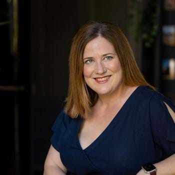 Sharon smiles at the camera. She has blue eyes, brown shoulder length hair, and is wearing red lipstick.