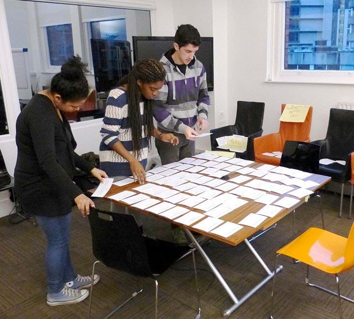 Two black women and one white man leaning over a table with cards spread over it.
