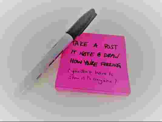 A Sharpie pen laid across the corner of a stack of pink post-its. Writing reads "Take a post it note & draw how you are feeling (you don't have to show anyone)"
