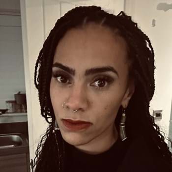 Kanika has braided hair and a nose ring stands in a room with soft lighting. She is wearing a dark top and earrings. They have a confident expression and are looking directly at the camera.