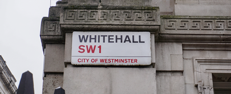 A sign reading "Whitehall SW1 is mounted on a white brick building, with the top of large gates seen at the bottom of the image