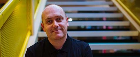 A smiling bald white man in a black t-shirt. He is sat on stairs with a yellow bannister.